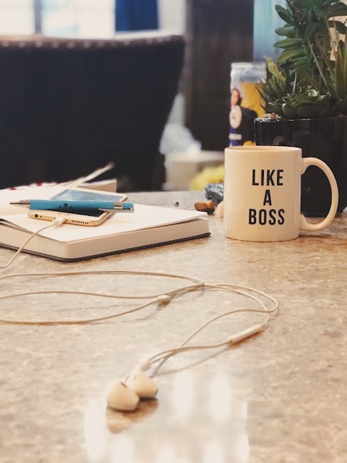 A close-up view of a cluttered table surface inside a home or office, with a white ceramic mug displaying the phrase 'LIKE A BOSS' positioned prominently in the center. To the left, there are a few closed notebooks and a smartphone resting on a white closed laptop. White earphone cables are loosely spread across the table, with the earbuds lying near the lower edge of the image. In the background, there is a potted plant with green foliage, a plastic bottle, and blurred elements of a room, suggesting an indoor environment that may be in the process of packing or organization, relevant to house removal or packing activities by Maidavale Removals.
