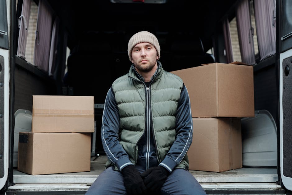 A man dressed in a beige knit beanie, dark gloves, and a green and navy padded jacket sits inside a moving van with the back doors wide open. Inside the van, several cardboard boxes of varying sizes are stacked on the floor, some placed on their sides, all sealed with packing tape. The interior of the van appears spacious, with dark fabric-lined walls and a neutral-colored floor designed for transporting household items. The scene is set outdoors, with the van situated on a street or driveway, and natural lighting illuminating the area. This image depicts the loading stage of a home relocation process, with furniture protection blankets and packing materials visible, aligning with the services offered by Maidavale Removals as featured on the webpage for Elgin Avenue man and van services for Maida Vale terraces.