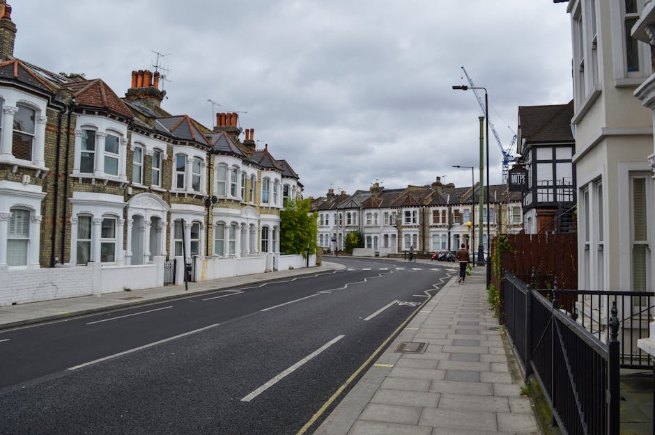 The image shows a quiet residential street in Maida Vale with a row of Victorian-style terraced houses featuring white and brick facades, bay windows, and multiple chimneys on rooftops. The street is paved with asphalt and marked with parking lines on one side, while the other side has a wide concrete sidewalk bordered by black metal railings. Several cardboard boxes, wrapped in plastic, and pieces of furniture are situated on the pavement near the entrance of one house, indicating a home relocation process. In the background, a man is seen walking away along the sidewalk, and a worker is loading or unloading items from a large white box truck with Maidavale Removals branding parked nearby. Construction cranes and street lights are visible above, and the sky is overcast with gray clouds, suggesting typical weather conditions for an urban move involving furniture transport and packing preparations that align with the services offered by Maidavale Removals for house removals in the Maida Vale area.
