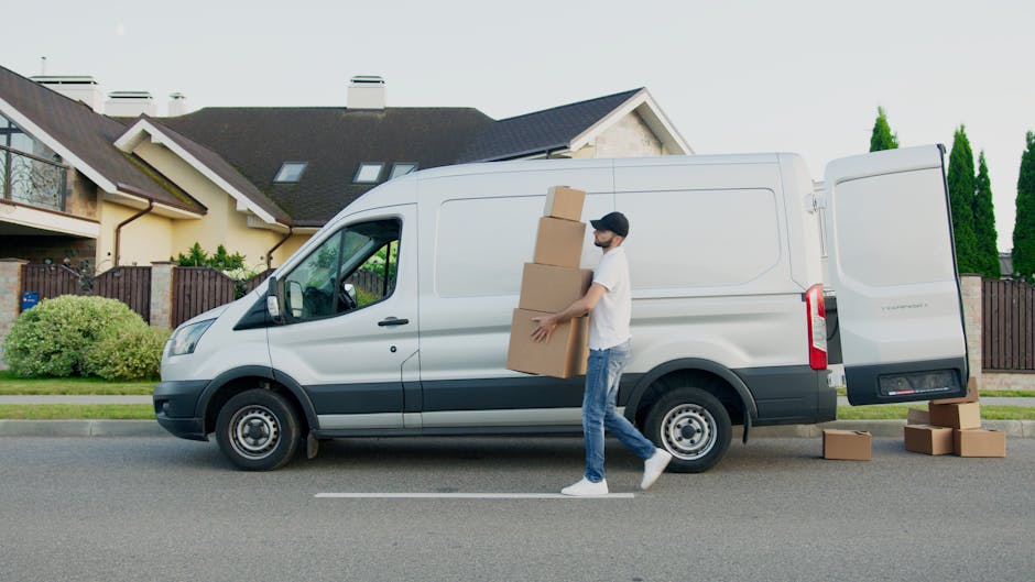 A man dressed in a beige knit beanie, dark gloves, and a green and navy padded jacket sits inside a moving van with the back doors wide open. Inside the van, several cardboard boxes of varying sizes are stacked on the floor, some placed on their sides, all sealed with packing tape. The interior of the van appears spacious, with dark fabric-lined walls and a neutral-colored floor designed for transporting household items. The scene is set outdoors, with the van situated on a street or driveway, and natural lighting illuminating the area. This image depicts the loading stage of a home relocation process, with furniture protection blankets and packing materials visible, aligning with the services offered by Maidavale Removals as featured on the webpage for Elgin Avenue man and van services for Maida Vale terraces.