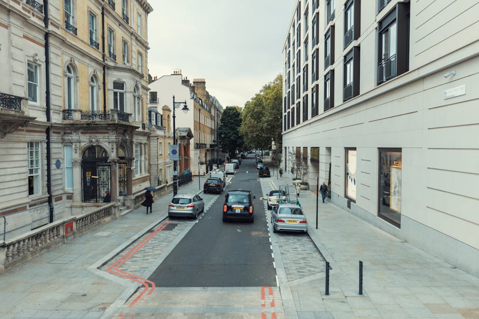 Photograph of a street in Westminster showing a row of elegant, multi-storey terraced houses with white facades, decorative black wrought iron balconies, and columns at the ground level. Several cars,