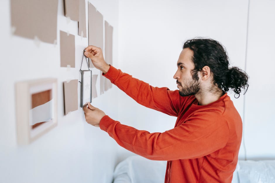 A man with long, curly dark hair tied back in a ponytail, wearing an orange long-sleeved shirt, is standing inside a home near a white wall decorated with several framed pictures and artworks. He is in the process of carefully wrapping or protecting the frames with transparent plastic film, likely as part of packing for a house move or furniture transport. He holds a roll of plastic wrap in his left hand and uses his right hand to stretch and smooth the material onto a frame. The scene suggests careful handling during packing and moving preparations, performed by Maidavale Removals, as part of their house removals and fragile item moving services. The lighting is natural and bright, indicating daytime, and the room appears tidy with a minimalistic aesthetic, emphasizing the focus on packing and moving activities related to home relocation.