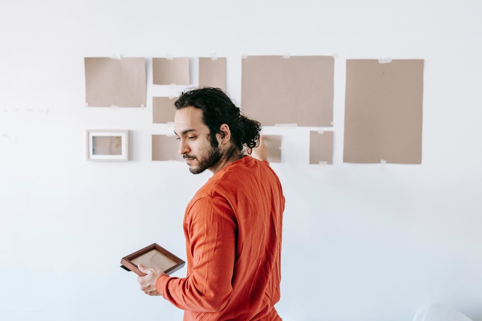 A man with dark, curly hair and a beard, wearing a red long-sleeve shirt, stands inside a room holding an open picture frame in his right hand and looking at a wall with multiple empty, beige picture mounts arranged in a grid pattern. The room has white walls and the mounts are set in a neat formation, suggesting preparations for hanging art or photographs. In the background, there are no visible furniture or packaging materials indicating a house removal or relocation process. The image captures a moment of organizing or decorating, with natural lighting highlighting the scene. Maidavale Removals, a professional house moving company, might use this visual to illustrate packing, handling, or furniture transport activities involved in home relocations, especially those involving art or delicate items. The setting shows an indoor environment during daylight, with a focus on the task of preparing or arranging artwork for moving or reinstallation, aligning with services like fragile move management and furniture transport for house removals.}