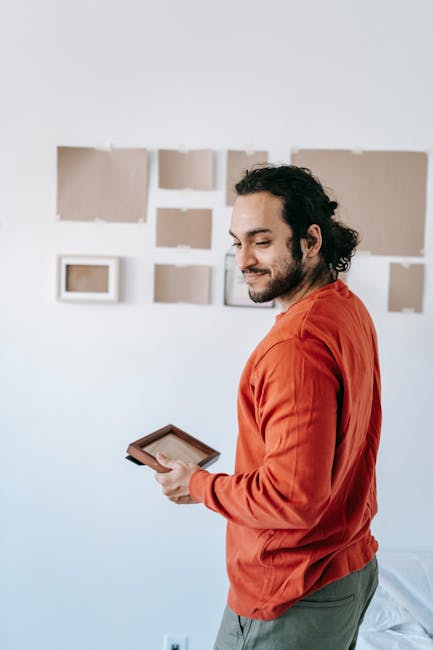 A man with long, curly dark hair tied back in a ponytail, wearing an orange long-sleeved shirt, is standing inside a home near a white wall decorated with several framed pictures and artworks. He is in the process of carefully wrapping or protecting the frames with transparent plastic film, likely as part of packing for a house move or furniture transport. He holds a roll of plastic wrap in his left hand and uses his right hand to stretch and smooth the material onto a frame. The scene suggests careful handling during packing and moving preparations, performed by Maidavale Removals, as part of their house removals and fragile item moving services. The lighting is natural and bright, indicating daytime, and the room appears tidy with a minimalistic aesthetic, emphasizing the focus on packing and moving activities related to home relocation.