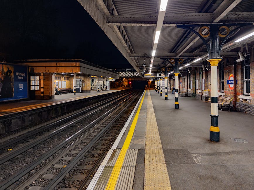 A view of an underground train station platform at night, showing multiple railway tracks with dark empty tunnels in the background. The platform is illuminated by overhead lighting and has yellow tactile paving strips along the edge for safety. Support columns with decorative yellow and black striped patterns are positioned at regular intervals along the platform. The station's brick exterior and the London Underground roundel sign indicate it is part of the UK rail network. The scene captures a quiet moment during a home relocation process, where moving boxes and packing materials are not visible, but the setting suggests the beginning or end of a furniture transport or moving operation is imminent, and Maidavale Removals could be engaged in coordinating the logistics of house removals in the Maida Vale area near the station W9.