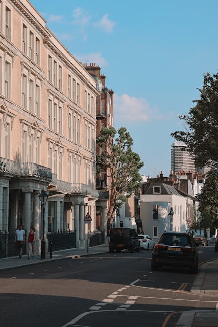 The image shows a residential street in Maida Vale with historic-style cream-colored terraced houses featuring sash windows, decorative cornices, and balconies. Parked cars line the curb, including a black van parked on the pavement, possibly used for home relocation or furniture transport. In the foreground, a loading area is visible where Maidavale Removals staff is engaged in a house removal process, with cardboard boxes, wrapped furniture, and packing materials arranged on the pavement near the vehicle. The scene is well-lit with natural daylight, and a large tree provides some shade. This setting highlights the logistics involved in packing and moving household items in an urban environment, supporting house removals services near Maida Vale station W9 by Maidavale Removals.