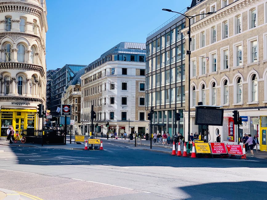 A daytime street scene in a densely built urban area with a mix of historic and modern architectural styles, featuring tall buildings with large windows and detailed facades. The street is partially closed off by temporary barriers and traffic cones, with signs indicating road closures and access restrictions. Pedestrians can be seen walking along the sidewalk, while a yellow and black vehicle, possibly a van used for house removals, is partially visible on the left side. In the foreground, there are various traffic management signs, some with yellow and red backgrounds, indicating access details. The lighting is natural, with clear blue skies overhead. This scene illustrates the logistical environment for house removals, with street access management and urban loading areas, relevant to the services offered by Maidavale Removals for house and office relocations, including furniture transport and packing activities.