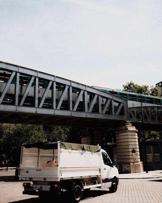 A white flatbed van, with its rear cargo area partially covered by a green tarp, is parked on a paved street beneath a metal railway bridge during daytime. The open flatbed contains cardboard boxes, packing materials, and wrapped items prepared for home relocation or furniture transport. The bridge, supported by large stone pillars, spans across the street, with a section of a train visible traveling above. Surrounding trees and buildings are visible in the background, indicating an urban area typical of Maida Vale. This scene captures the loading process involved in professional removals or packing and moving services, with careful positioning of boxes and equipment ready for transportation, illustrating the logistical steps undertaken by Maidavale Removals during same day or urgent relocation projects.