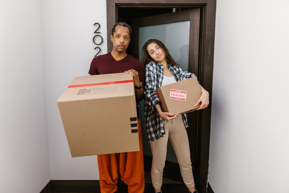 A man and woman stand inside a residential entrance doorway during a home relocation process, each holding cardboard boxes filled with household items. The man, dressed in an orange pair of trousers and a maroon top, holds a large box in front of him, while the woman, wearing beige trousers and a checked shirt, holds a medium-sized box marked with a 'Fragile' label. Behind them, the interior wall is painted white, and the dark wooden doorframe surrounds a gray door, partly open, revealing a corridor or interior space. The scene is lit with natural indoor lighting, capturing the moment of packing and moving, with the boxes likely prepared for transport by Maidavale Removals or similar home removals services. This snapshot illustrates the packing and loading process involved in house removals, emphasizing the careful handling of household goods during a same day move in Maida Vale, as part of professional furniture transport or relocation services.
