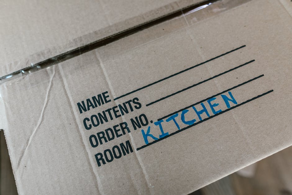 Close-up of a cardboard moving box with printed labels in black and blue ink, indicating contents and room designation. The box is taped with clear packaging tape and features handwritten text in blue marker specifying 'KITCHEN' as the designated room. It appears to be part of home relocation or packing and moving procedures carried out by Maidavale Removals. The box is placed inside a property, likely on the floor or a trolley, ready for loading or transport, with a focus on packing materials and organization during the furniture transport process. The environment suggests an indoor setting, with neutral lighting highlighting the details of the packing label and the cardboard texture, supporting the process of house removals and storage management.