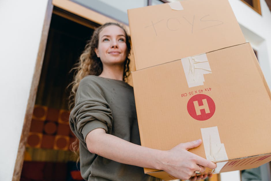 A woman with long, curly hair and light skin is standing inside a home near a doorway, holding a large cardboard box labeled 'toys' and a smaller box. She is dressed in a grey long-sleeve top with rolled-up sleeves and has a gentle, content expression on her face. The larger box has white packing tape and a red circular sticker with an 'H' on it, indicating it is part of a home relocation packing process. In the background, there are audio speakers or decorative items, and natural light illuminates the scene, suggesting daylight inside the property. The image captures the active process of packing and loading items in preparation for house removals, a task often managed by professional moving services such as Maidavale Removals, who offer assistance with furniture transport and moving logistics as seen in this context.