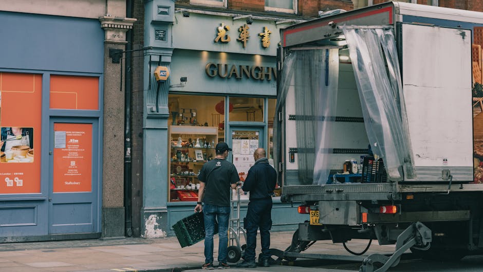 Two movers from Maidavale Removals are preparing for home relocation outside a building with a Chinese-themed storefront, featuring gold Chinese characters and the word 'GUANGH.' One mover, dressed in dark clothing, is carrying a green plastic crate, while the other, in a black jacket, is handling a hand truck. A large, open delivery truck with a partially covered cargo area is parked on the pavement, ready for furniture transport and packing materials. Inside the truck, visible packaging supplies include plastic wrap and cardboard boxes, indicating staging for a professional moving process. The scene captures the loading process in an urban setting, emphasizing the preparation involved in house removals, with the company's services supporting efficient packing, furniture transport, and logistics for property relocations in the Maida Vale area. The environment is well-lit with natural daylight, highlighting the equipment used in the move and the building’s exterior details.