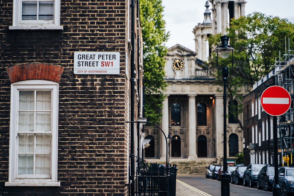 A street scene in Westminster showing the corner of a brick building with a white-framed window and a street sign for Great Peter Street SW1 mounted on the wall. In the background, there is a classical-style building with columns, a clock, and steps leading up to its entrance. A black street lamp and a red no-entry sign are also visible, along with parked cars lining the street. The image captures an urban environment typical of Westminster, with elements relevant to house and furniture removals, such as the street's accessibility and urban logistics for relocation services by Maidavale Removals.